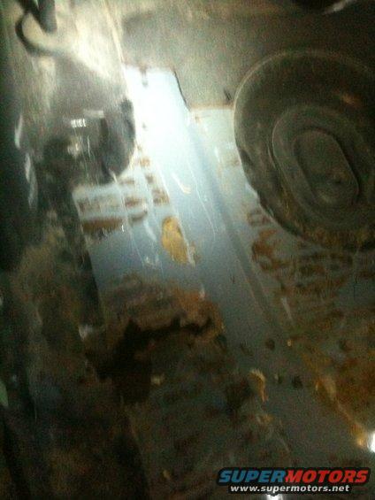 Undercarriage of a vehicle with significant rust and damage. A tire is partially visible on the right