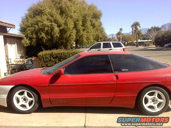 1995 Ford Probe GT coupe, side view, red with white wheels. White SUV parked behind it