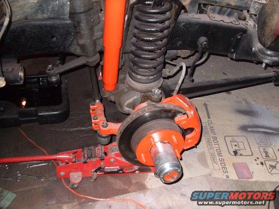 Vehicle front suspension with orange shock absorber, brake caliper, and rotor assembly, supported by a red hydraulic jack