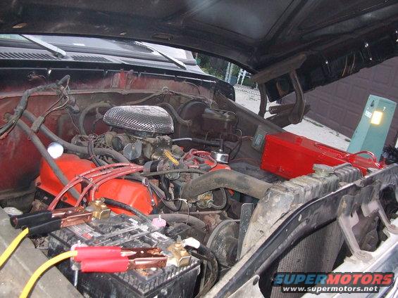 Engine bay of a red 1978 Ford Bronco with jumper cables attached to the battery and an open hood