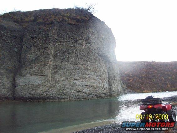 Red ATV with black cargo box on rear, parked on gravel shore next to water