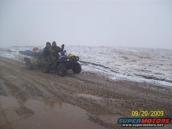 Two people ride an ATV that is being towed by a truck in a snowy, muddy landscape. The truck bed is loaded with gear