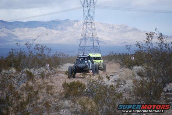 Two off-road race buggies driving on a dirt track with mountains and a power tower in the background