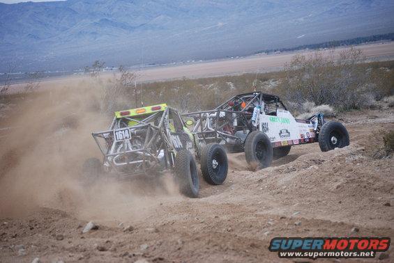 Two off-road buggies kicking up dust on a dirt track, one with number 1614