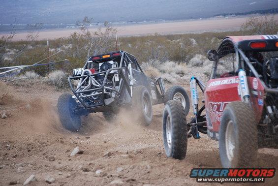 Two off-road buggies kicking up dust on a dirt track, one black with number 1695, the other red