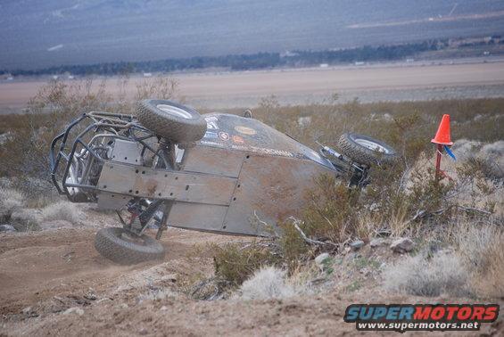 Rolled-over off-road buggy in desert brush, tires visible, roll cage, dirt terrain
