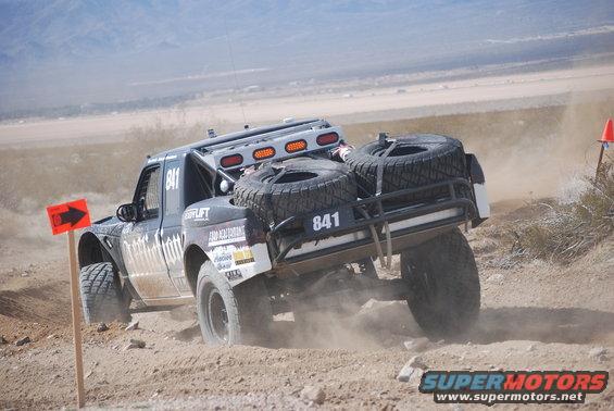Off-road racing truck with number 841, carrying two spare tires on the rear rack, kicking up dust on a dirt track