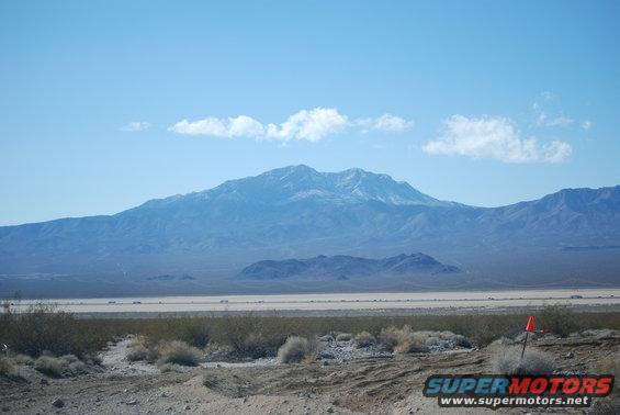 Desert landscape with mountains under a blue sky. Orange marker visible in foreground