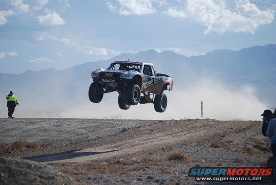 Off-road truck mid-air jump, dust cloud behind it. Person in yellow vest watches