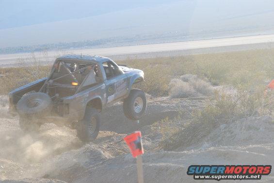 Off-road truck mid-air, kicking up dust, with a spare tire mounted on the rear