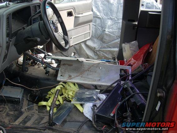 Interior of a vehicle with the dashboard removed, showing exposed wiring, tools, and various parts scattered around