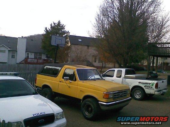 Yellow Ford Bronco with a black soft top, parked next to a white pickup truck and a police car
