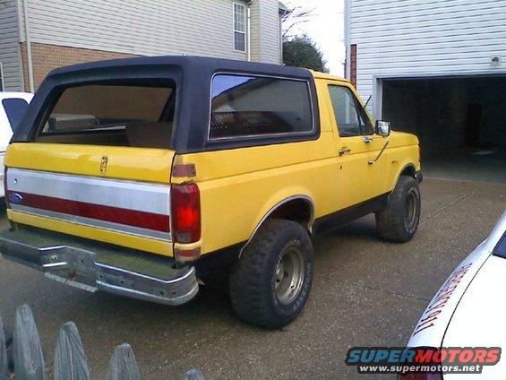 Yellow Ford Bronco parked in driveway, rear view. Has a black removable top and chrome bumper
