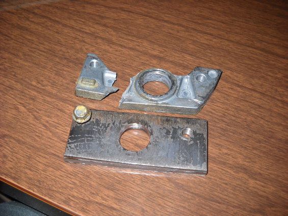Broken metal bracket with a circular opening and a separate metal plate with a bolt. These parts are on a wooden surface