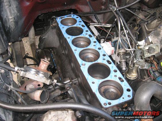 Engine block with a blue gasket installed over the cylinders. Various engine components and hoses surround the block