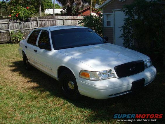 White 2002 Ford Crown Victoria parked on grass in front of a wooden fence and a shed