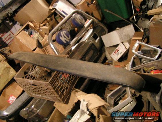 Pile of car parts including a bumper, KC lights, and various trim pieces, with boxes and a milk crate