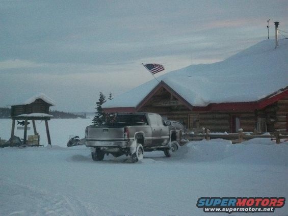 Silver pickup truck parked in snow in front of a log cabin with an American flag
