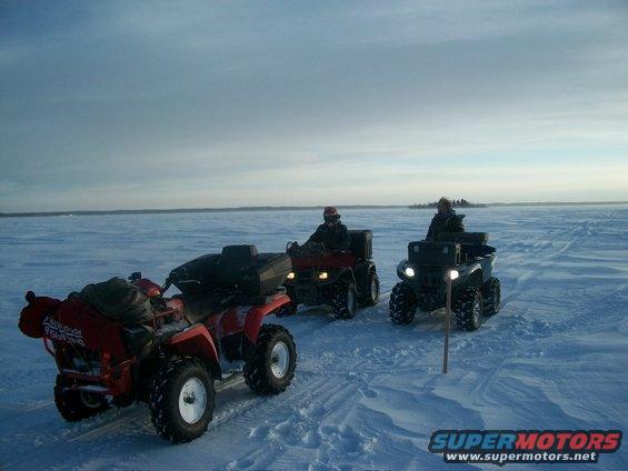 Three ATVs ride across a wide, flat expanse of ice under a pale blue sky. Two riders are visible