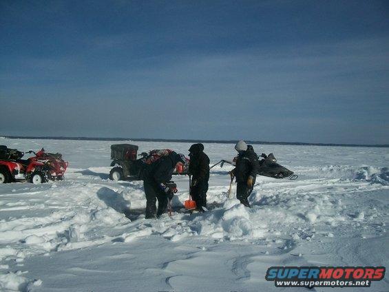Two red ATVs and one dark ATV with a cargo box sit on a snow-covered frozen lake with people drilling holes