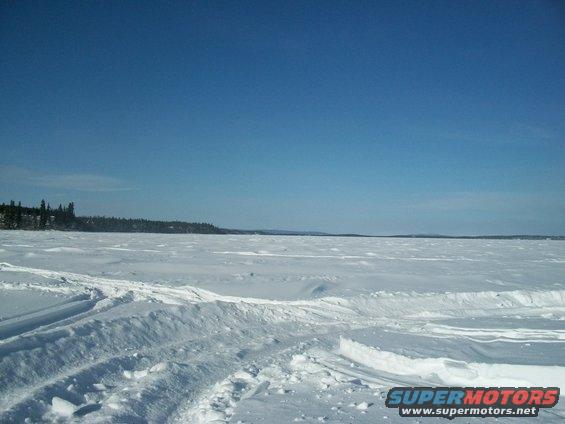 Wide expanse of snow with tire tracks leading into the distance under a clear blue sky