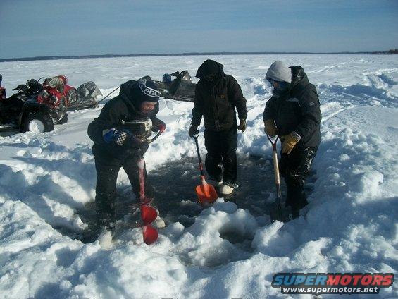 Three people are ice fishing on a frozen lake, one using an auger. ATVs and sleds are parked nearby