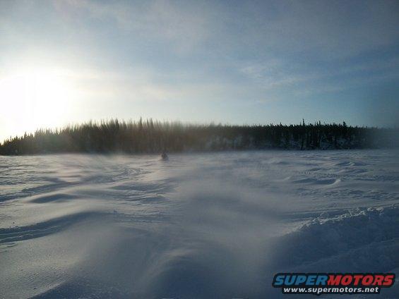 Snowmobile moving across a snowy field with trees in the background and sun low in the sky