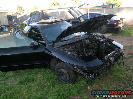 Black coupe with hood and driver door open, engine exposed, cardboard on roof, parked on grass. Another black coupe visible behind