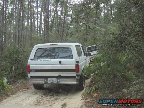 White Ford Bronco driving on a dirt road in a wooded area. Another vehicle is visible behind it