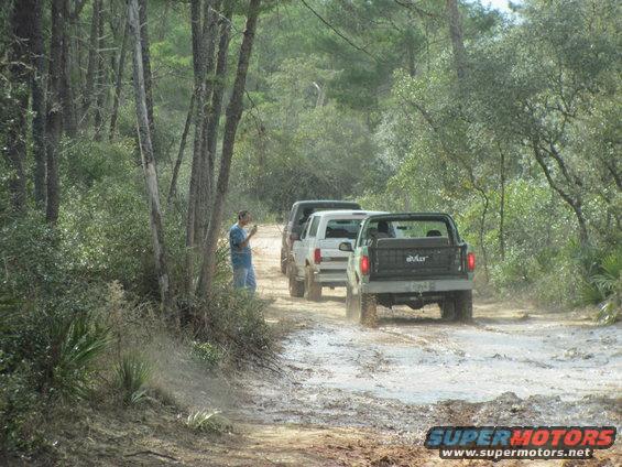 Three SUVs driving on a muddy dirt road, one is a 1990 Ford Bronco