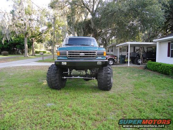 Teal lifted Ford truck with oversized tires, front view. Other vehicles visible