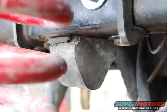 Close-up of a cracked rubber body mount attached to a metal bracket, with red components in the foreground. Supermotors.net logo visible