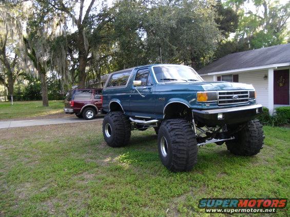 Teal lifted Ford Bronco with huge tires parked in a grassy yard. Another SUV is parked behind it