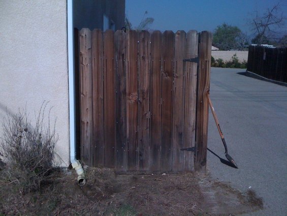 Wooden gate with a shovel leaning against it. Building on the left, asphalt driveway on the right