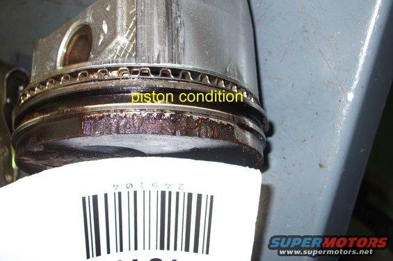 Engine piston with carbon buildup and visible wear. A barcode is below the piston