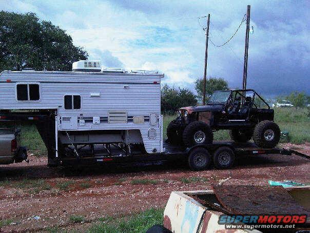 Off-road vehicle with roll cage on a trailer, next to a camper. Supermotors.net logo visible