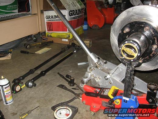 Automotive steering and brake components laid out on a garage floor with tools and a jack