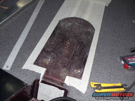 Template taped to a dark, textured surface, possibly for a boat or vehicle floor part. Utility knife and tape measure nearby