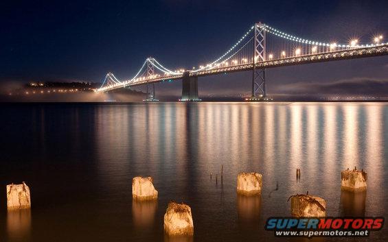 Bay Bridge illuminated at night with reflections on the water. Some old pilings are in the foreground