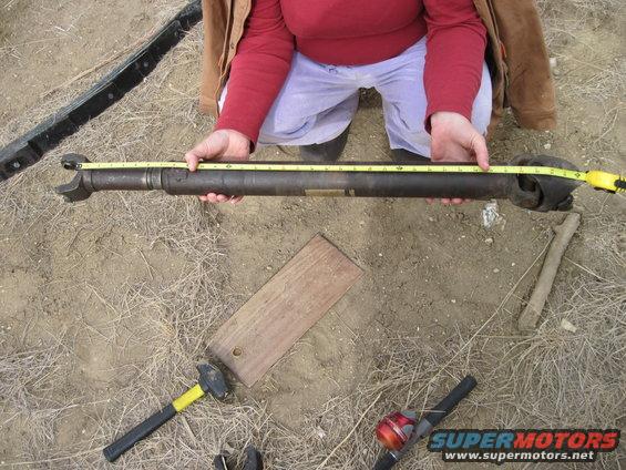 Person measuring a driveshaft with a yellow tape measure. Hammer and wood plank on the ground