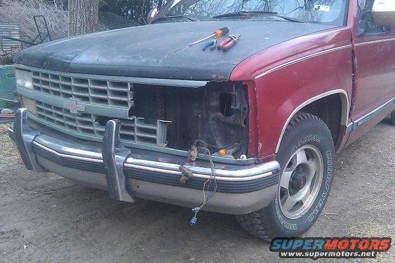Red pickup truck front end with missing headlight and grille, tools on hood