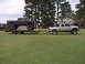 1985 Ford Bronco II - A black lifted pickup truck is transported on a trailer pulled by a silver lifted pickup truck across a grassy field.