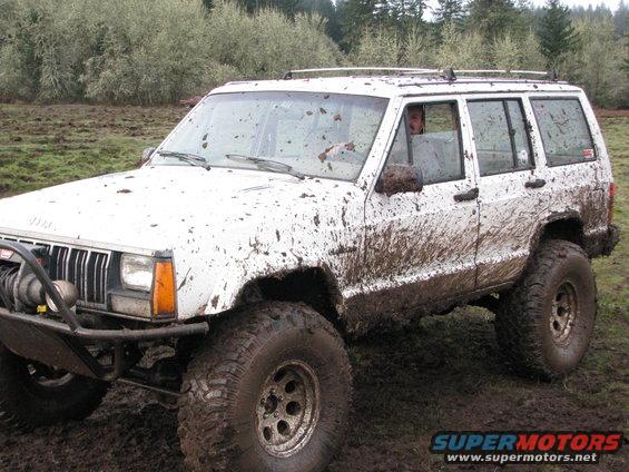 White Jeep Cherokee with large off-road tires, covered in mud, driving through a muddy field