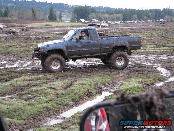 Muddy blue pickup truck with large tires in a mud bog, driver visible