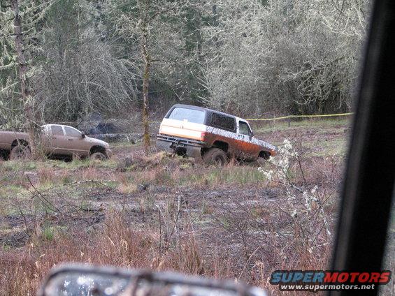 Two muddy trucks stuck in a wooded area. One is a white and orange SUV, the other is a tan pickup