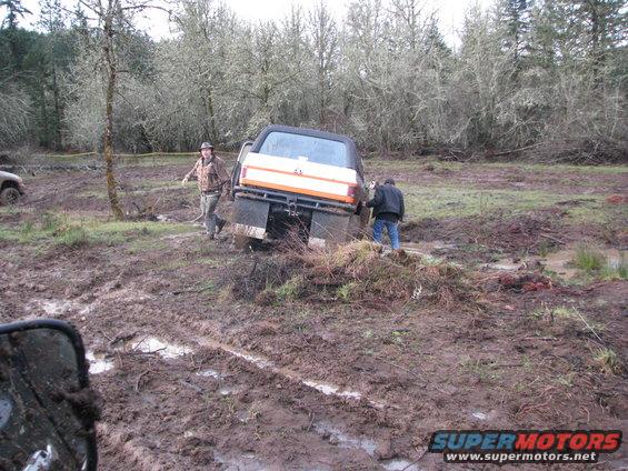 Orange and white pickup truck with dual rear wheels stuck in mud, viewed from the rear