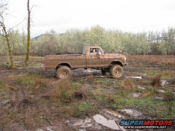 Muddy pickup truck with lifted suspension driving through a wet, grassy field with trees in the background