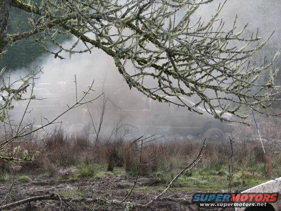 SUV partially obscured by smoke, side view, muddy terrain, branches in foreground
