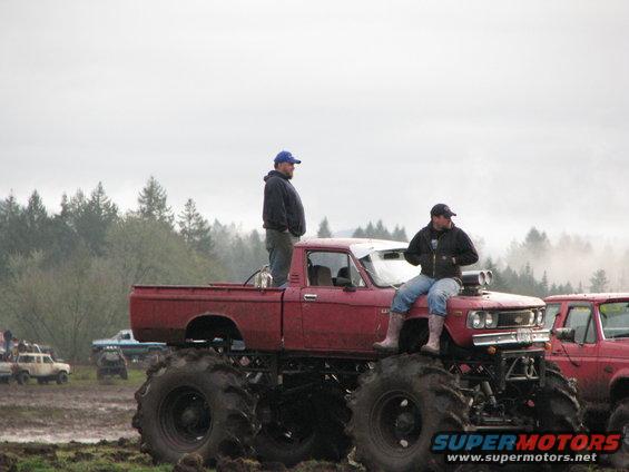 Red monster truck with large tires and two men, one standing in the bed and one sitting on the hood
