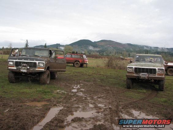 Two Ford F-Series trucks in a muddy field, one brown and one green. A red SUV is in the background